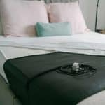 A rectangular black Earthing Bed Mat - Universal lying across a white bed sheet, with a coiled black lead and a plug resting on its surface.