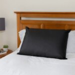 A black Earthing Pillow Cover rests on a pillow against a wooden headboard on a white bed. To the left, a bedside table holds a lamp and small plant.