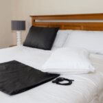 A neatly made bed with white sheets, pillows, a black mat, and an Earthing Pillow Cover with a coiled cord on top. A bedside table with a lamp is visible in the background.