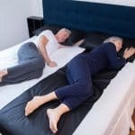 An older man and woman sleep comfortably on a large bed, separated by a wide black Earthing Elite Universal Sleep Mat. Both wear pajamas and appear relaxed, each resting on their own side of the bed.
