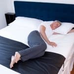 A man rests peacefully on his side on a bed with white sheets, a dark headboard, and an Earthing Elite Universal Sleep Mat featuring a black strip across the lower part of the mattress, his eyes closed.