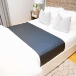 A neatly made bed with white sheets, two light gray pillows, and a dark gray runner features an Earthing Elite Universal Sleep Mat. A nightstand with a lamp and flowers sits beside the woven headboard and light rug.