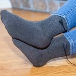 A person wearing Grounding Socks with a power cord attached sits on a wooden floor, legs extended and crossed at the ankles, wearing blue jeans.