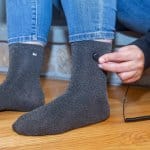 Someone in blue jeans sits on a wooden floor, plugging a small cord into the port of their gray Grounding Socks.