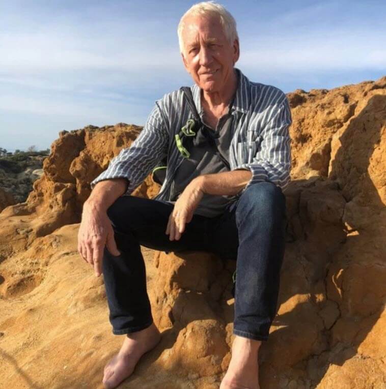 Clint Ober sitting barefoot on rocky, orange terrain, wearing a striped shirt over a gray t-shirt and jeans. He looks at the camera with a slight smile under a blue sky.