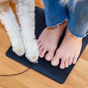 A person in frayed blue jeans and a white, curly-furred dog stand together on a Mini Universal Earthing Mat, which is black and rectangular, placed on a wooden floor.