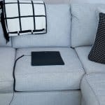 A Mini Universal Earthing Mat with a cord is placed on the seat of a light gray sofa, along with a white and black checkered blanket and a black-and-white patterned cushion.
