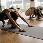 A group practices yoga indoors on Grounders Yoga Mats, stretching forward in childs pose as natural light streams through large windows in the background.