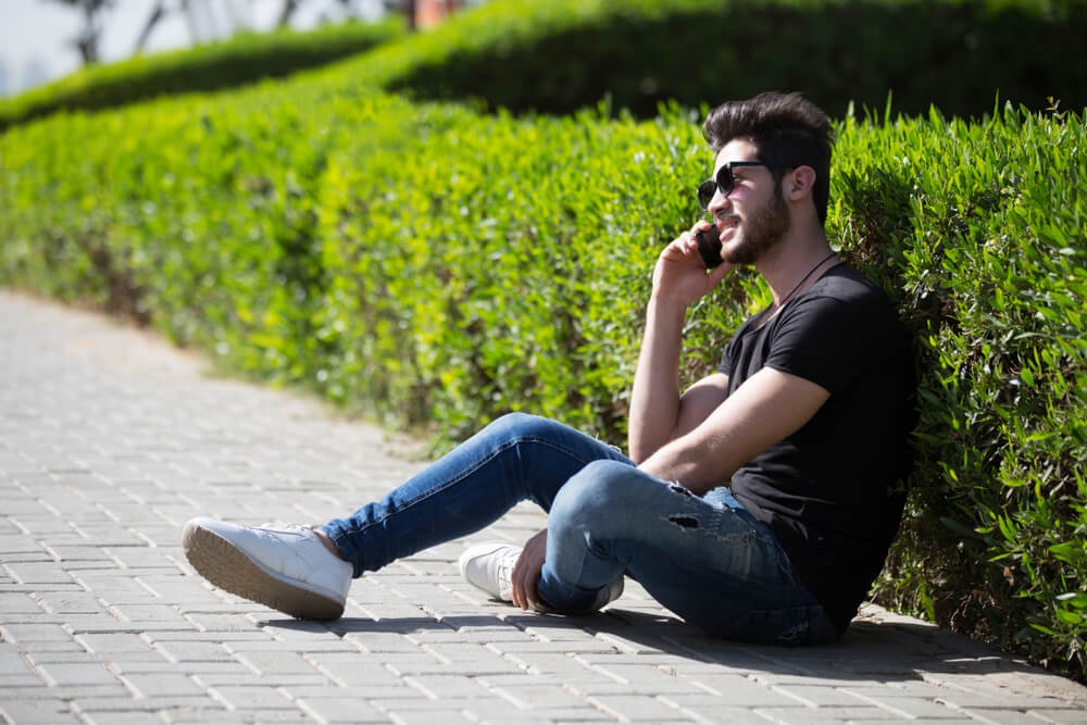 young guy in jeans with sunglasses leaning against bush