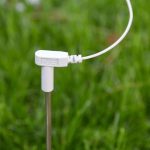 An Earthing Grounding Rod, a white electronic soil sensor with an attached cable, is inserted into green grass outdoors.