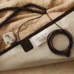 A close-up of a beige earthing mattress cover with a tag, black cords, and a UK plug adapter, all laid out on a textured tan surface.