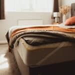 A neatly made bed with gray sheets, an orange blanket, and a mattress protector visible under the bedding in a sunlit bedroom with a window, gray curtains, and a nightstand with a lamp and vase.