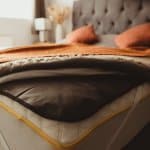 A close-up of an unmade bed, showing a mattress with a yellow trim, a gray weighted blanket, and an orange throw. The bed has a tufted headboard and two orange pillows, with soft natural light in the room.