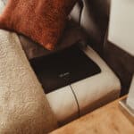 A black electronic device with a cord is placed under a brown pillow and beige blanket on a bed, next to a nightstand.