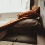 A neatly made bed with earth-toned sheets and pillows. A dark rectangular device labeled earthing with a cord is partially visible under the blanket, suggesting grounding or earthing technology in use.