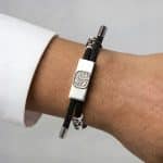 An Earthband PEMF Bracelet on a wrist, featuring a black cord, a rectangular silver plate with a tree-like design, and two silver accent beads.