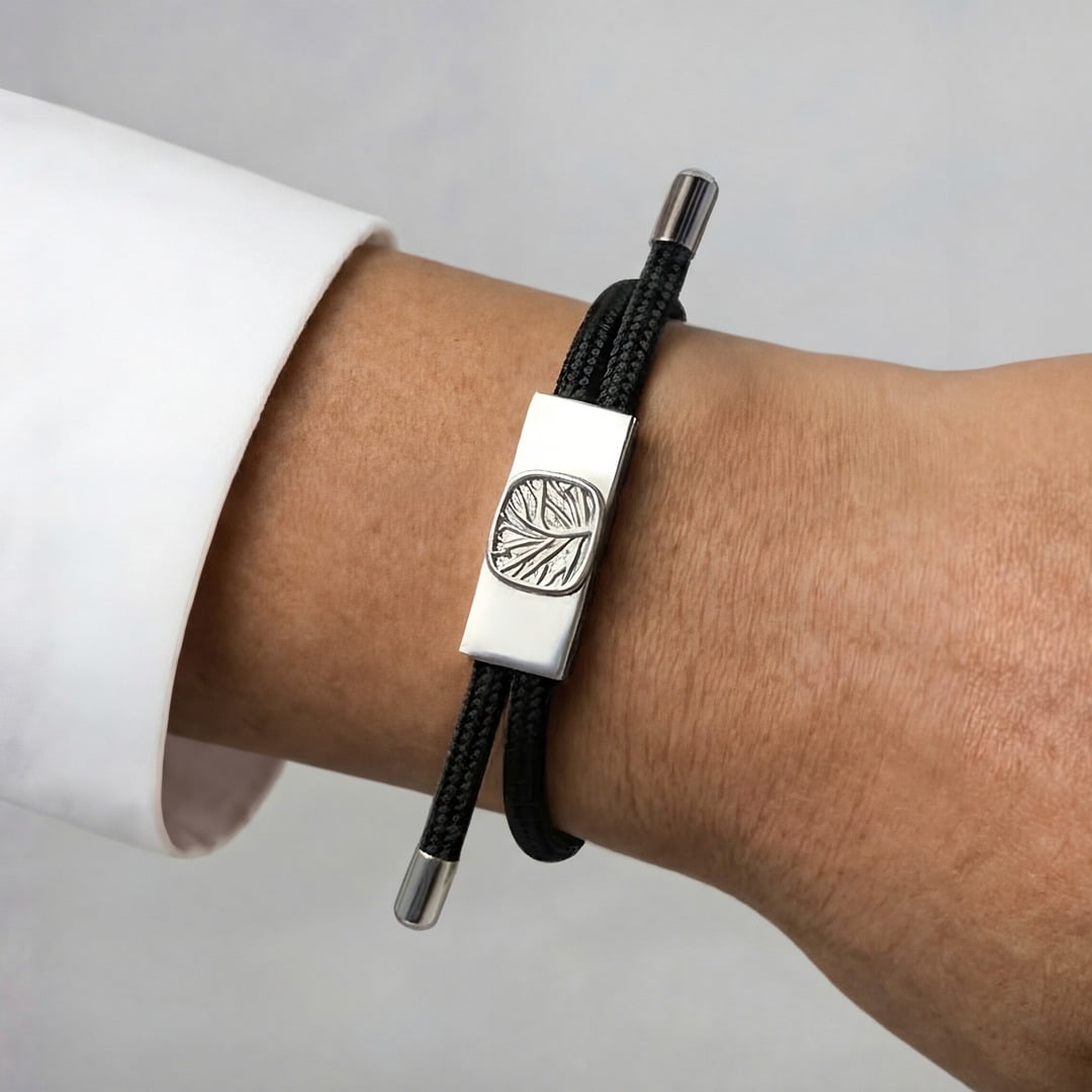 Earthband PEMF Bracelet featuring a rectangular silver-tone metal charm with an embossed leaf design on a black cord, worn on a wrist.
