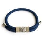 The EarthBand PEMF Bracelet from BeGrounded, featuring an engraved leaf design and a metal clasp on a blue cord, is elegantly coiled in a circle on a plain white background—showcasing its natural elegance in simplicity.