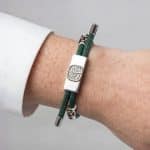 An Earthband PEMF Bracelet worn on a wrist, featuring a green cord, a rectangular silver centrepiece with an embossed tree design, and two silver metal accents.