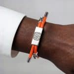 An orange Earthband PEMF Bracelet on a wrist, featuring a silver rectangular centrepiece with an embossed tree design and silver end caps.