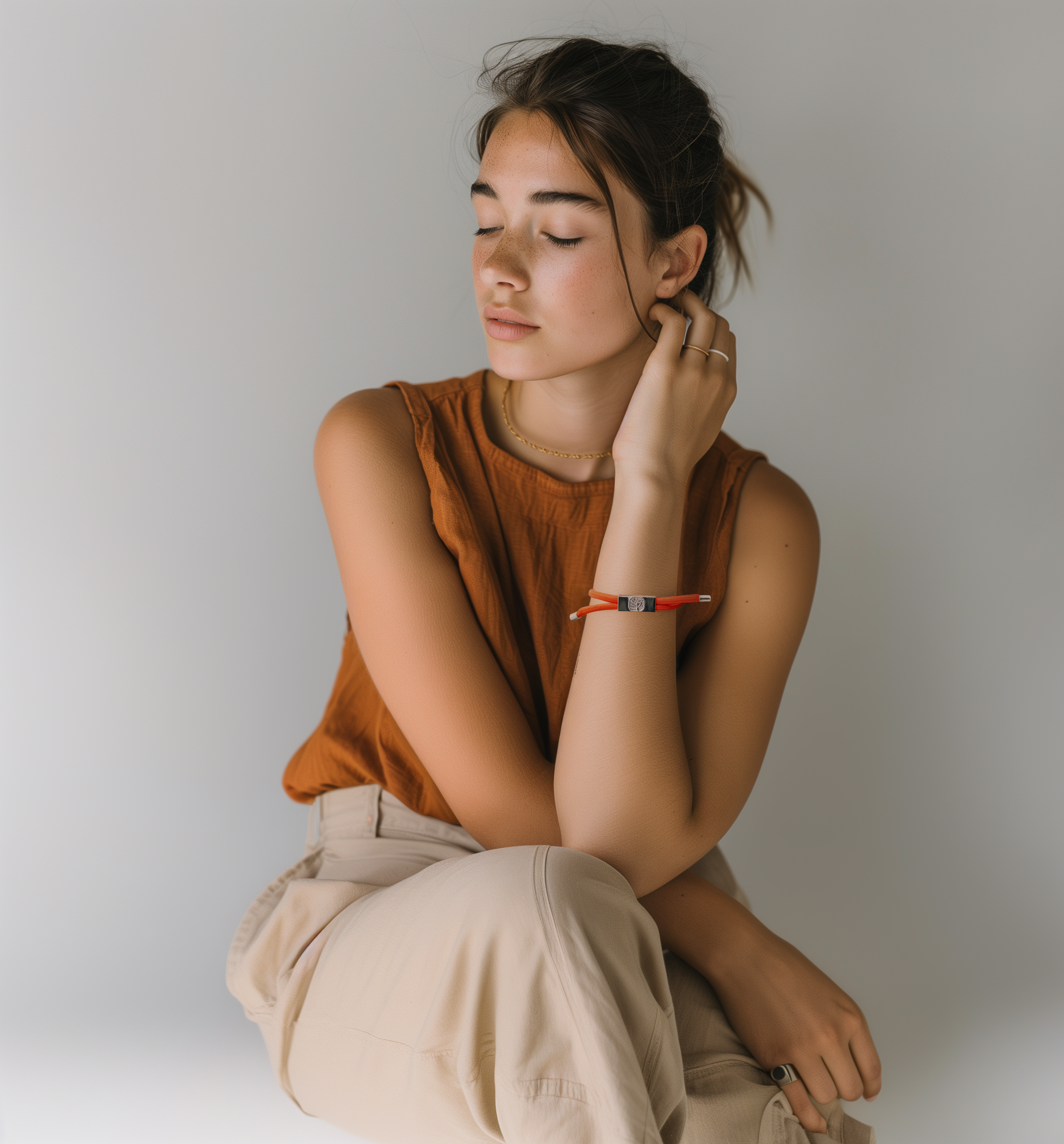 A young woman with closed eyes poses calmly against a plain background, wearing a sleeveless burnt orange top, light beige pants, and the Earthband PEMF Bracelet in red, appearing relaxed as if enjoying its soothing effects.