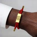 An Earthband PEMF Bracelet featuring a red cord and a rectangular gold-coloured charm engraved with a tree design, worn on a wrist.