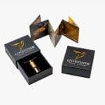 A gold and silver Lovetuner pendant on a chain sits in an open black box with gold logo and text. Next to it is another closed box and an unfolded brochure with images and text.