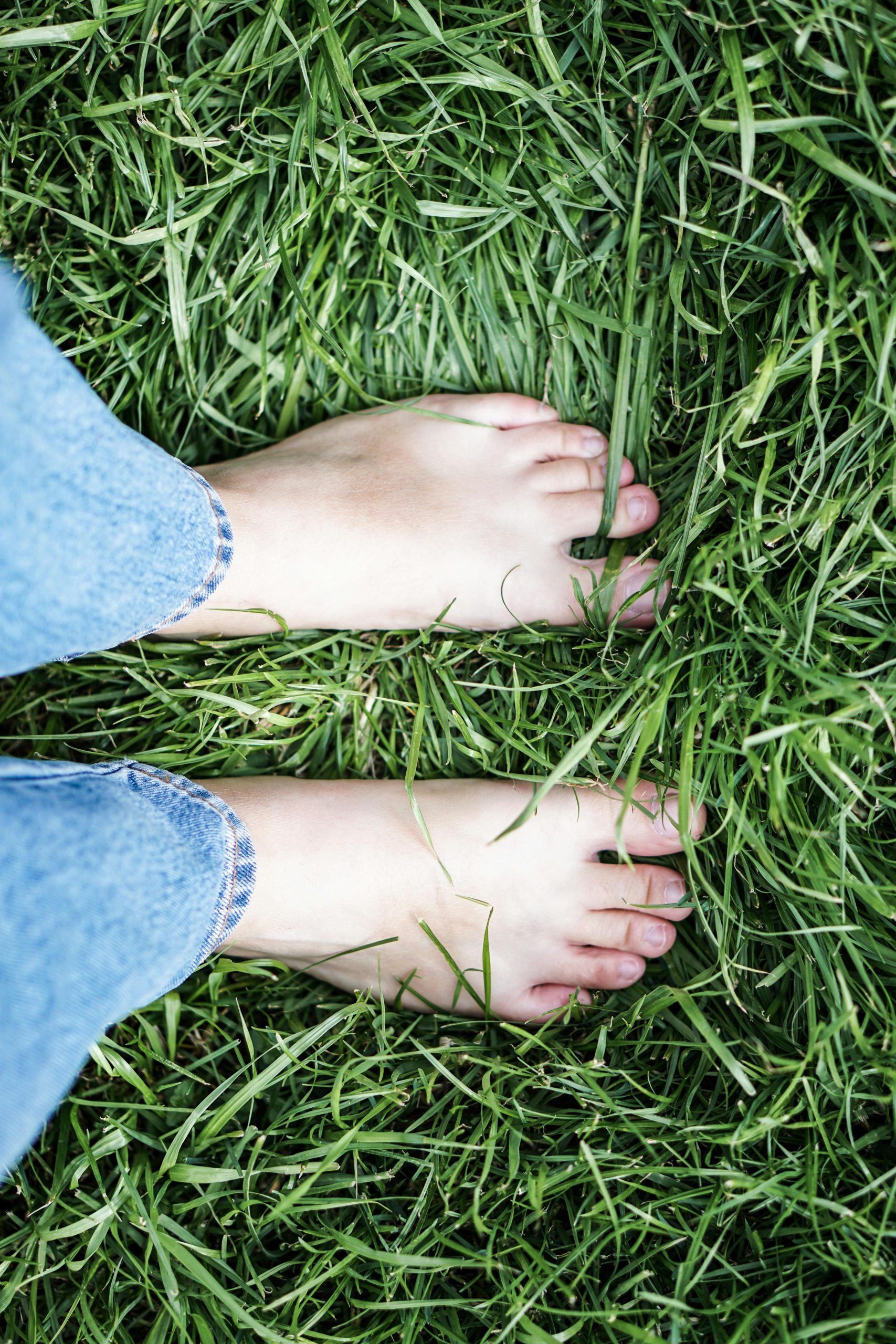 Bare feet standing on lush green grass, with the person wearing light blue rolled-up jeans—a peaceful moment showing How To Stay Grounded. The tall, healthy grass and parallel feet create a sense of calm connection to nature.