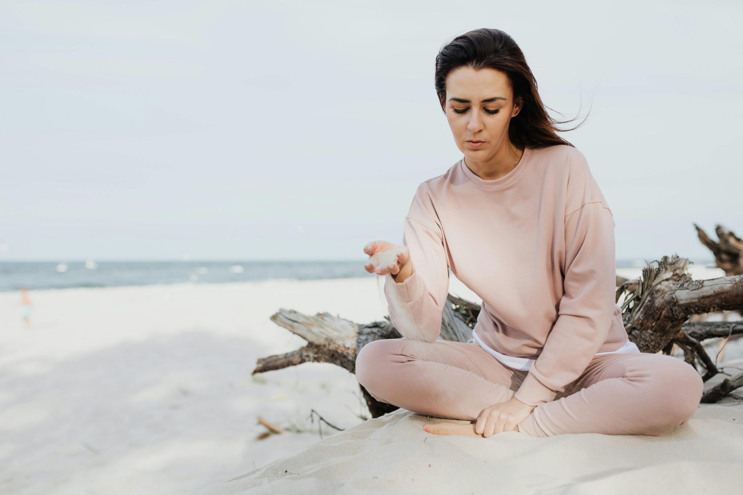 A woman in a light pink outfit sits cross-legged on sand near driftwood at the beach, looking down thoughtfully with her hand slightly raised—an ideal setting for Portable Grounding Solutions for Travelling, with the ocean and sky as her backdrop.