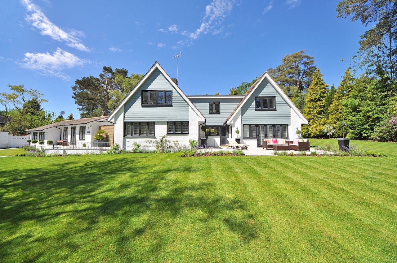 A modern house with gray siding and white trim features large windows and a well-manicured green lawn, surrounded by trees and shrubs under a partly cloudy blue sky, illustrating the importance of safety features like grounding in home design.