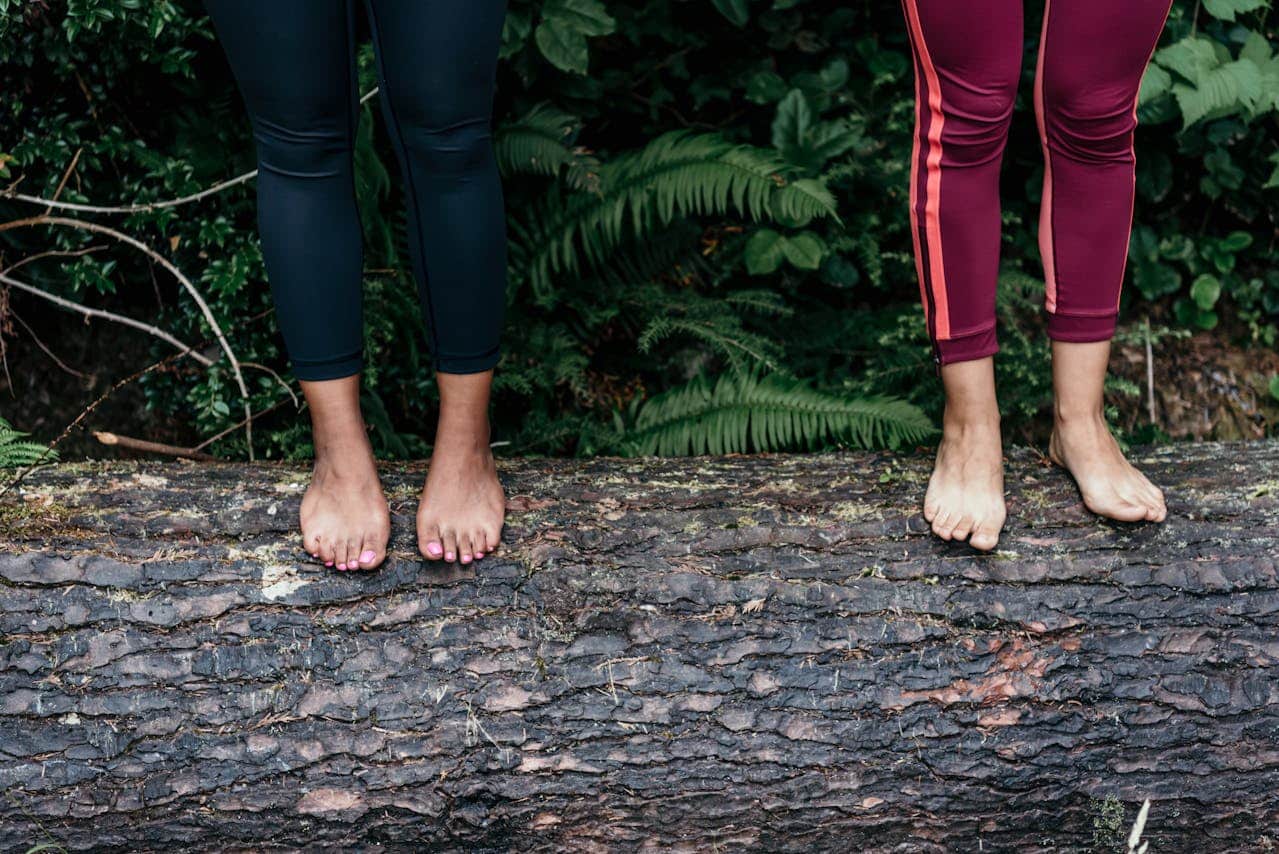 Two people in leggings stand barefoot on a large fallen tree trunk, surrounded by green foliage—a peaceful scene that evokes the calm of Anxiety Grounding Exercises. Only their legs and feet are visible.
