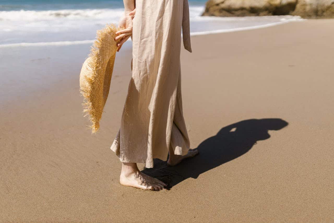 A person in a beige dress walks barefoot on a sandy beach while holding a straw hat, embracing the natural connection often linked to earthing. Ocean waves and rocks are visible in the background.