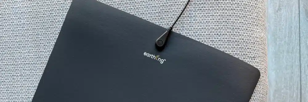 A black Earthing® mat with a connecting cord rests on a textured light gray fabric surface, next to a wooden floor—perfectly complementing your Grounding Sheet for an enhanced grounding experience.