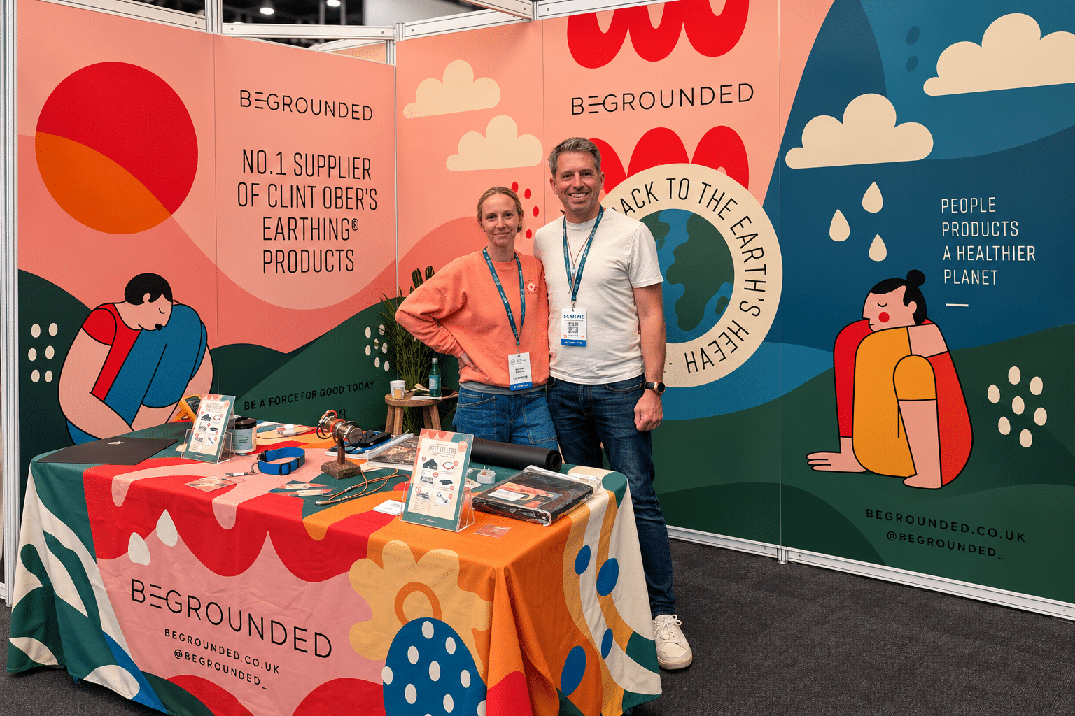 Two people stand smiling at a colourful Be Grounded stall with bold, abstract earth-themed graphics, promoting earthing products. The table displays items, signs, and business cards, with vibrant artwork on the backdrop and tablecloth. BeGrounded exhibits at The Health Optimisation Summit, sharing their innovative offerings in an engaging and visually striking space.