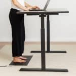 A person stands at an adjustable height desk typing on a laptop, with a flat heating mat placed on the floor under their bare feet.