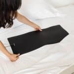 A person laying a rectangular black heating mat onto white bed sheets.