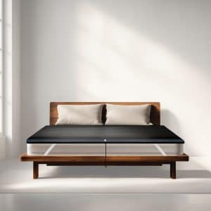 A mattress on a wooden bed frame covered by a dark, thin topper with a power lead hanging from the centre.