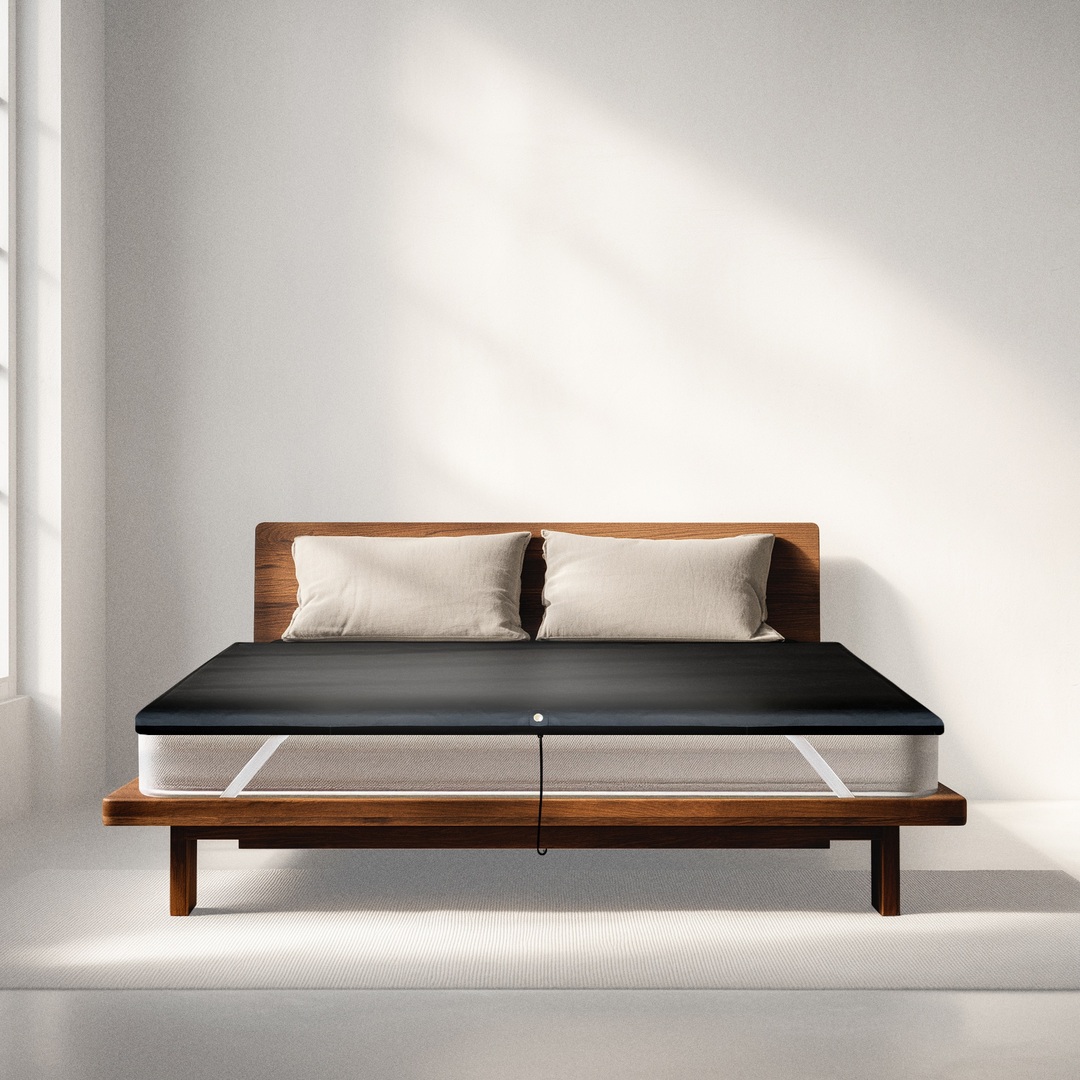 A mattress on a wooden bed frame covered by a dark, thin topper with a power lead hanging from the centre.