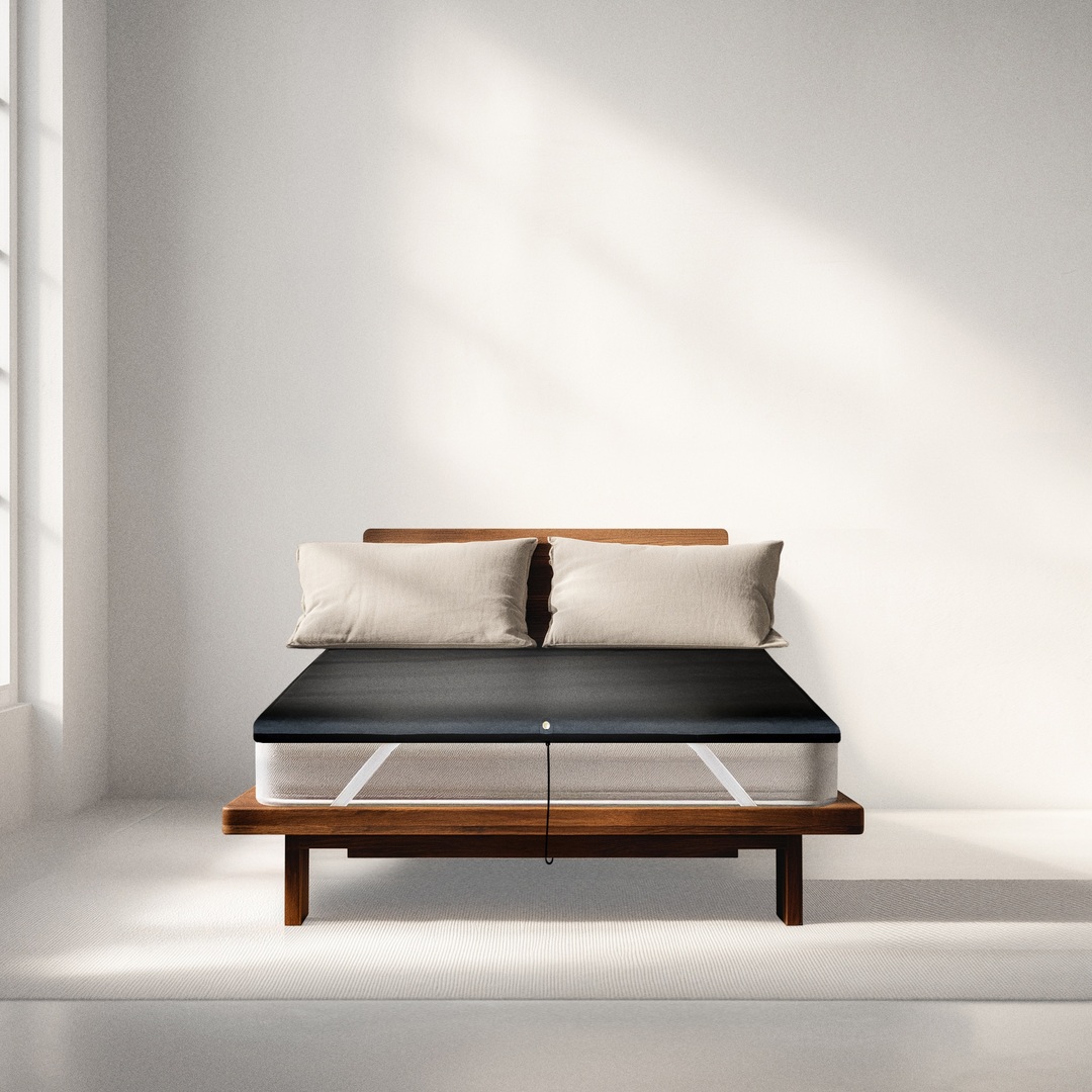 A black mattress topper with a power lead resting on a bed frame with two pillows.