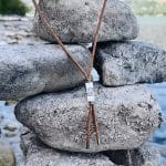 An Earthband PEMF Necklace with a tan cord and a rectangular silver bead featuring an etched leaf design, displayed resting against a stack of smooth grey stones.